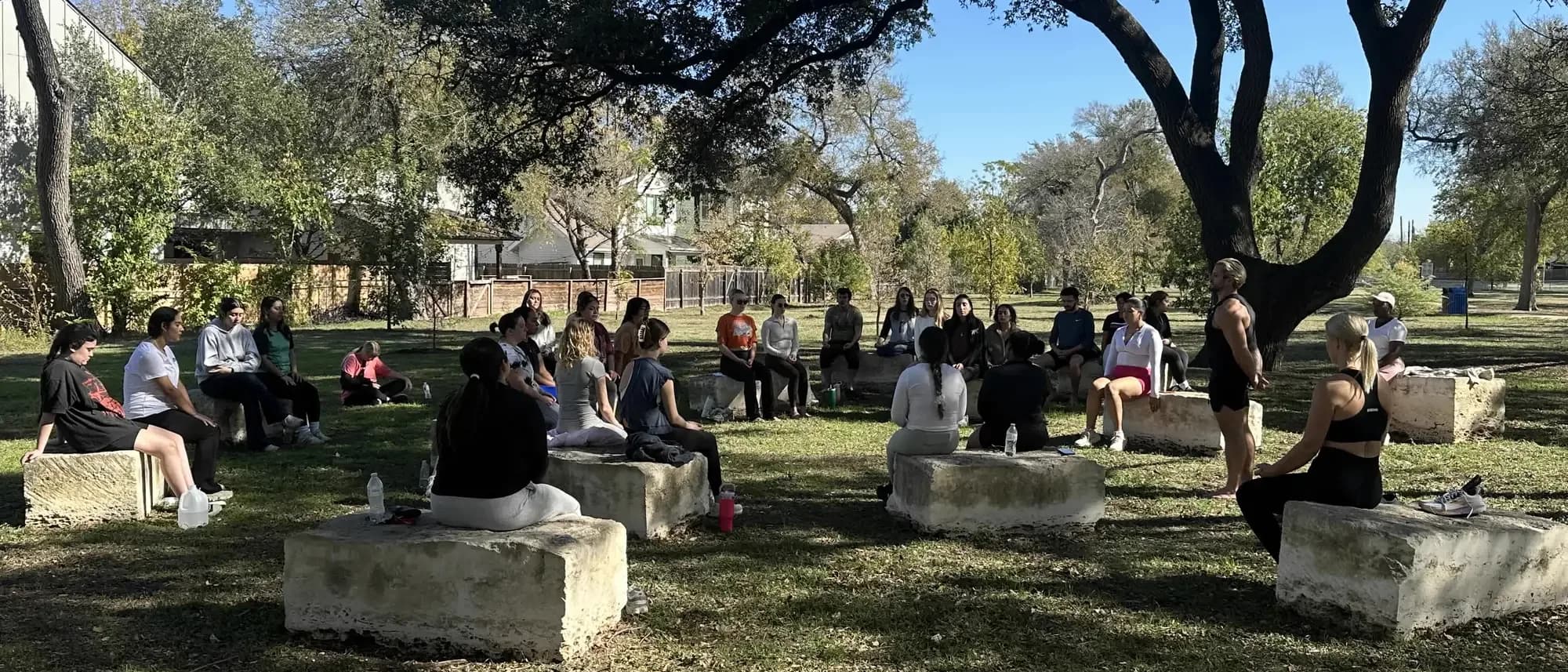 Jarred Curcio leading a group seated in a circle on the stone blocks under the oak trees in Austin, Texas